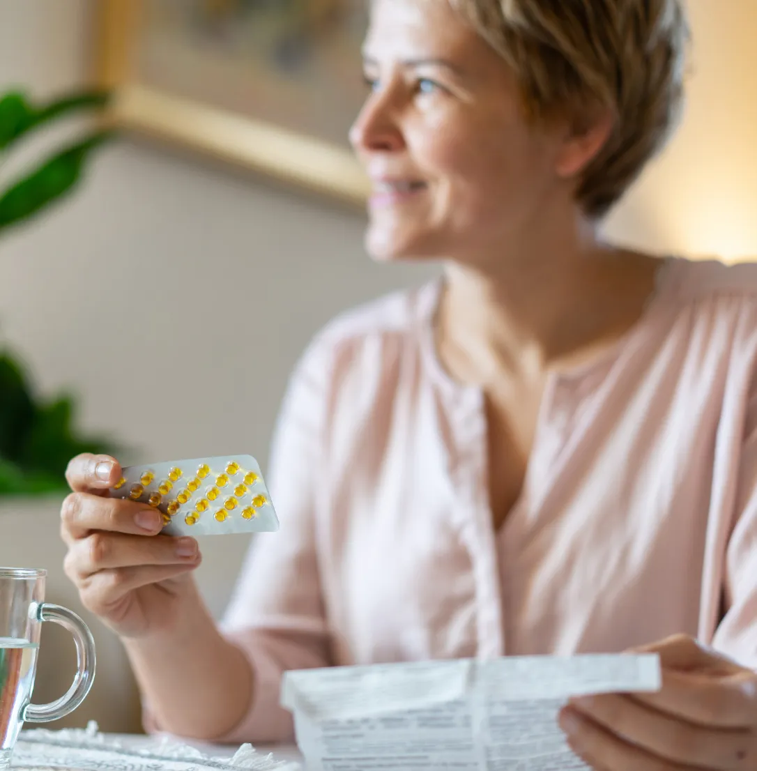 Person holding a blister pack of pills and a folded instruction leaflet, with a glass of water on the table.