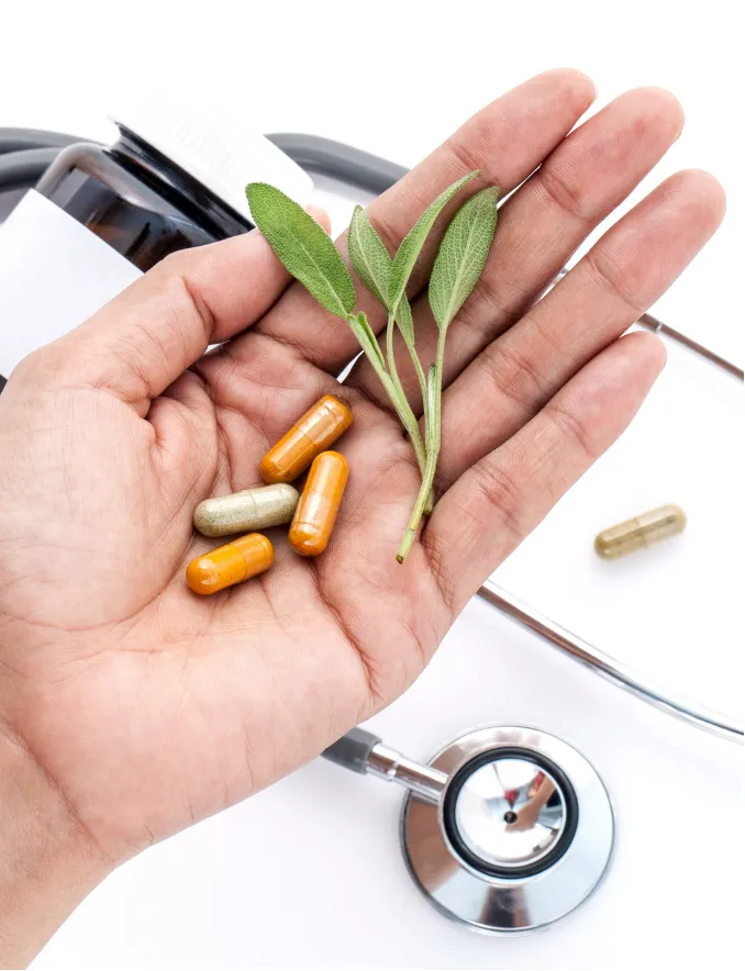 Hand holding capsules and leaves, with a stethoscope and pill bottle in the background on a white surface.