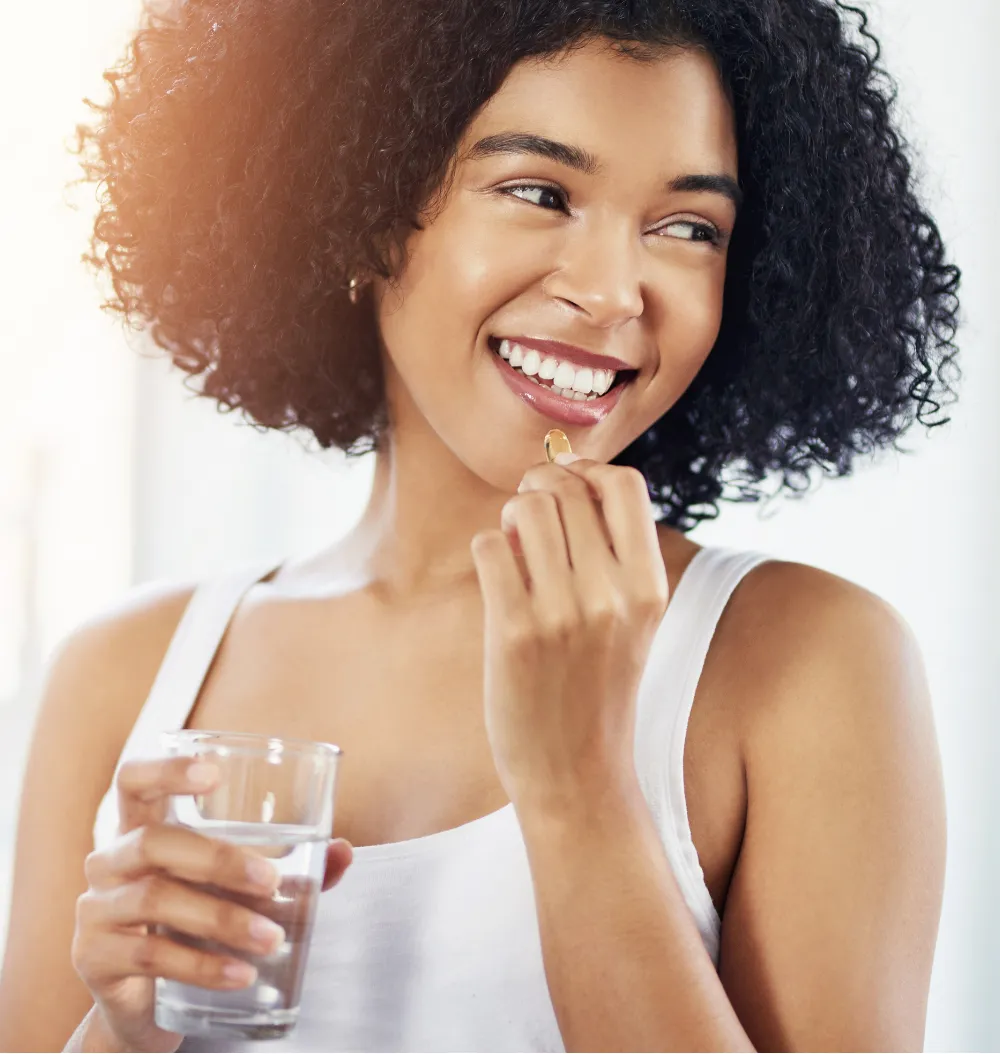 Woman with curly hair smiles while holding a pill and a glass of water.