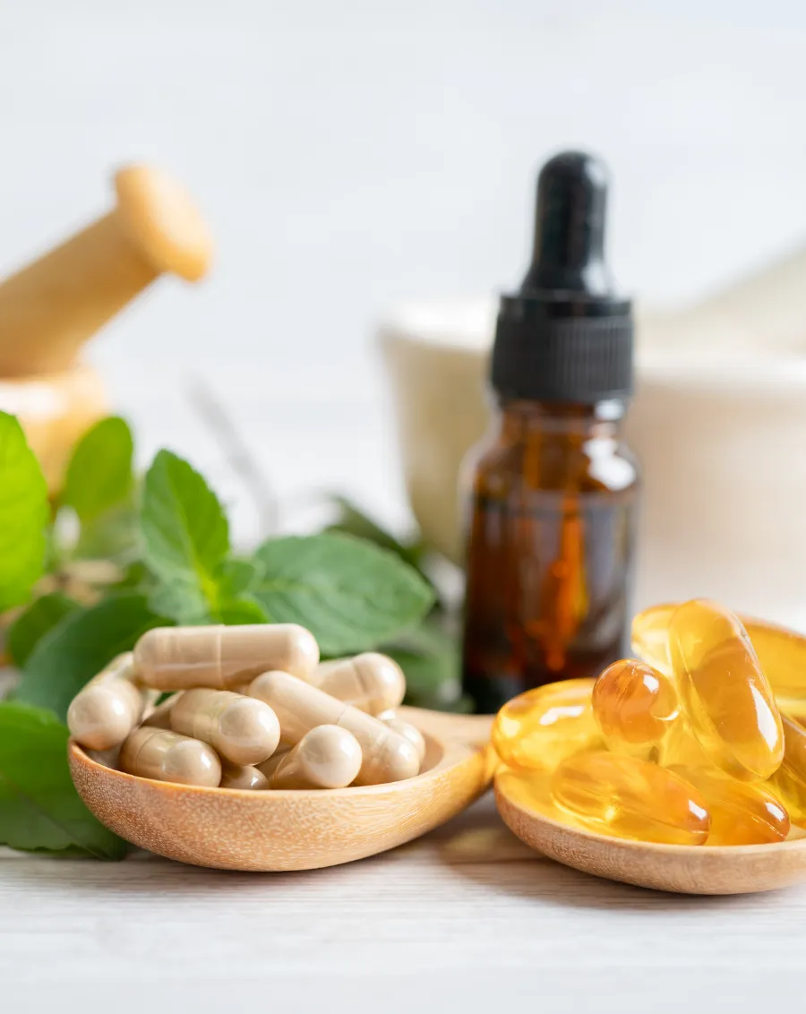 Wooden spoons with capsules and gel pills, dropper bottle, leaves, and a mortar in the background on a white surface.