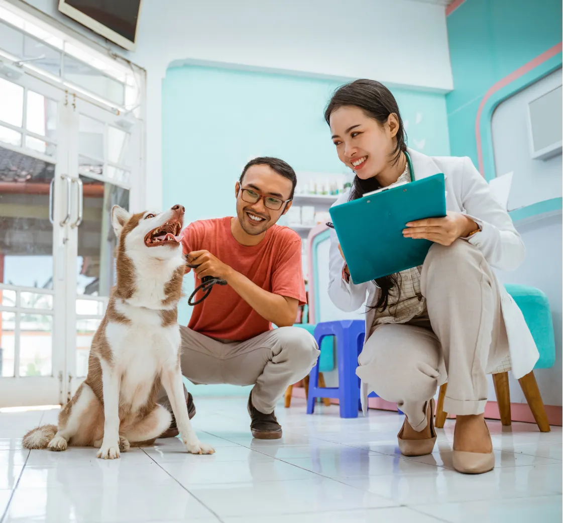 A happy dog sits inside a vet clinic with a smiling man and a vet holding a clipboard.