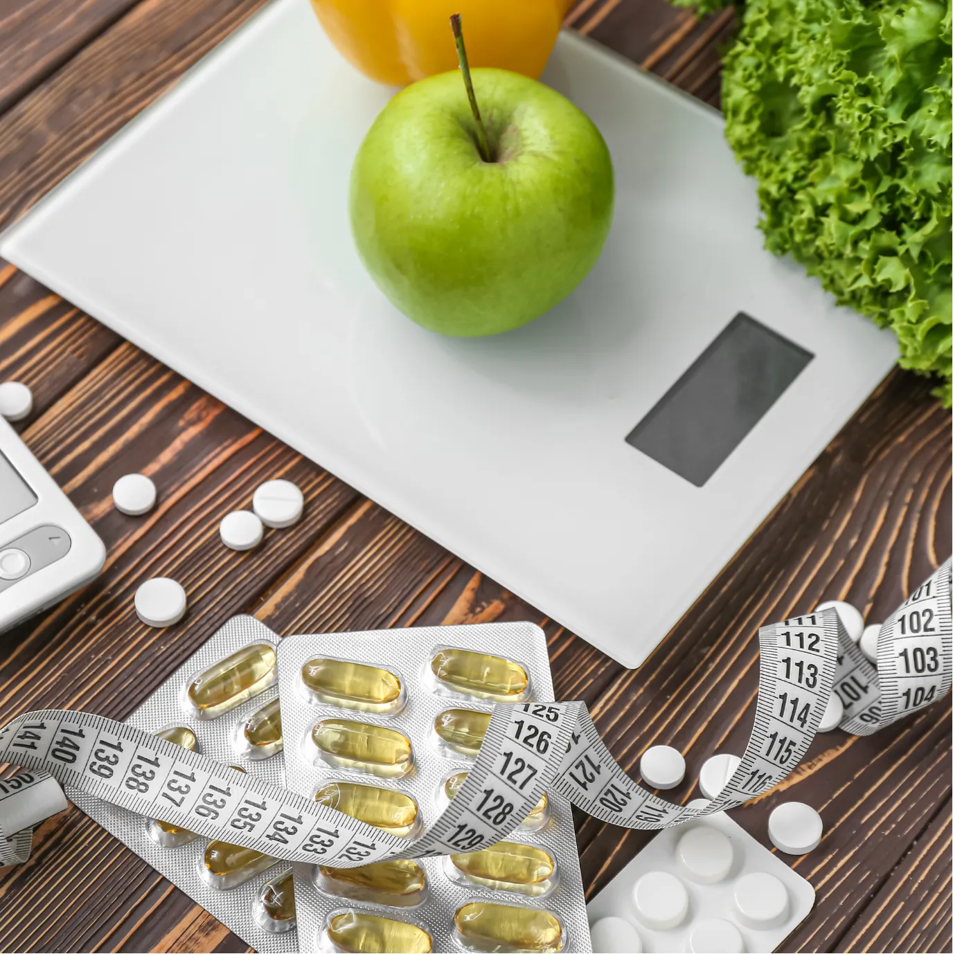 Digital scale with apple, pills, and measuring tape on wooden table, surrounded by lettuce and light orange beverage.