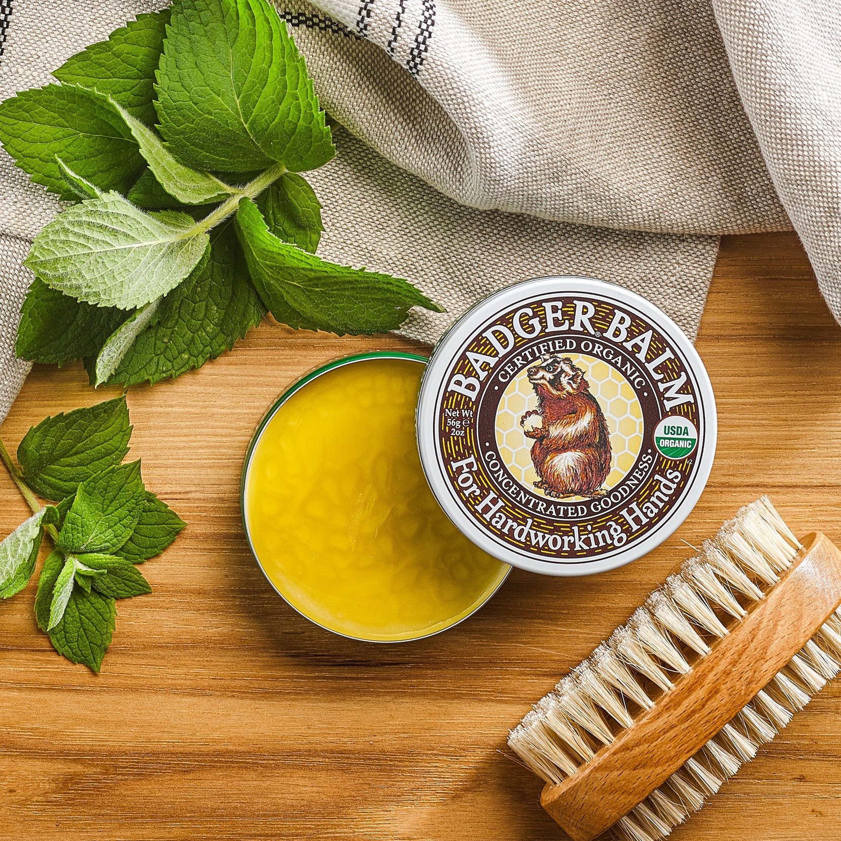 Open tin of yellow balm with mint leaves and a brush on a wooden surface.