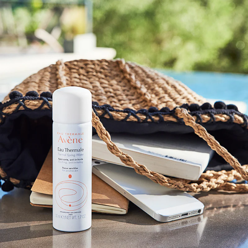 A bottle of Avène Eau Thermale spray next to a woven bag, books, and a smartphone by a pool.
