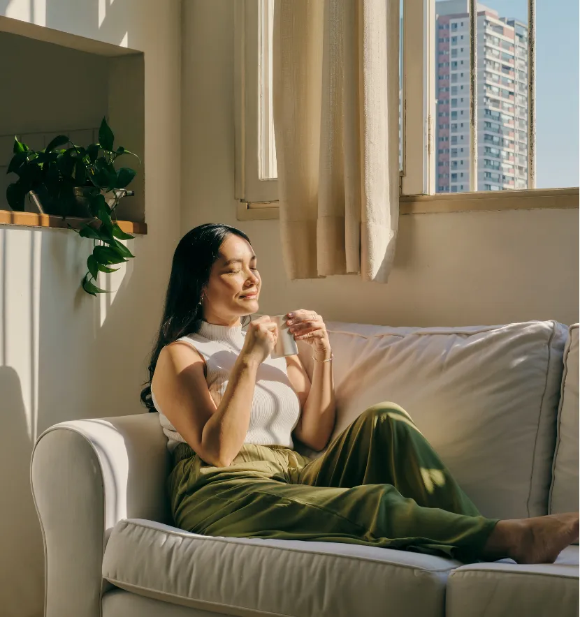 Woman relaxing on a couch, holding a coffee cup, with sunlight streaming through a window.