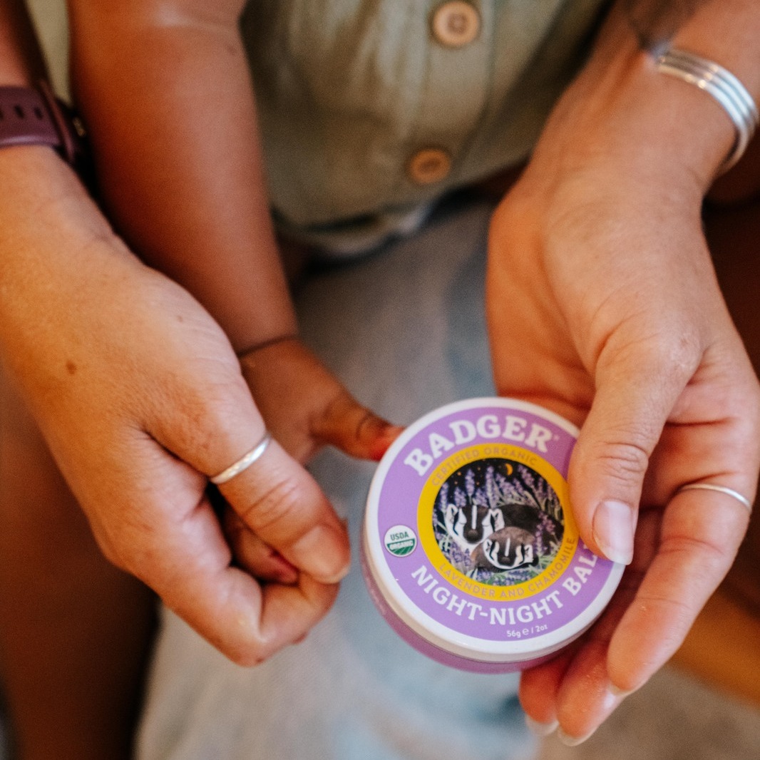 Hands holding a small container of Badger Night-Night Balm with a purple lid.