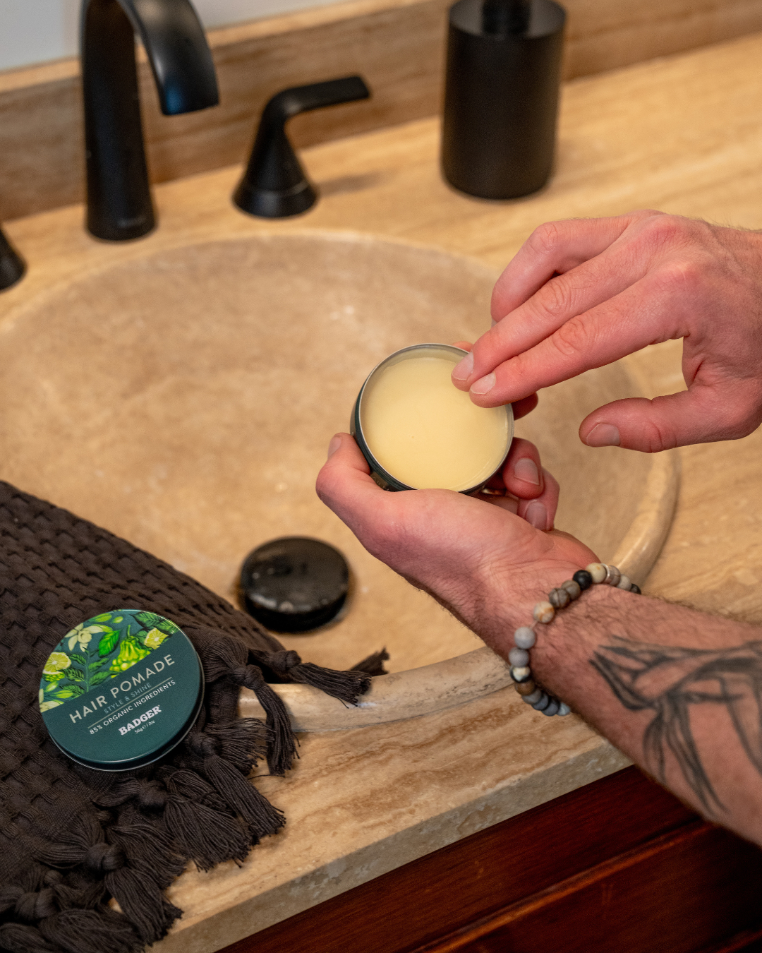 Person scooping hair pomade from a container, near a bathroom sink with a black faucet and towel.