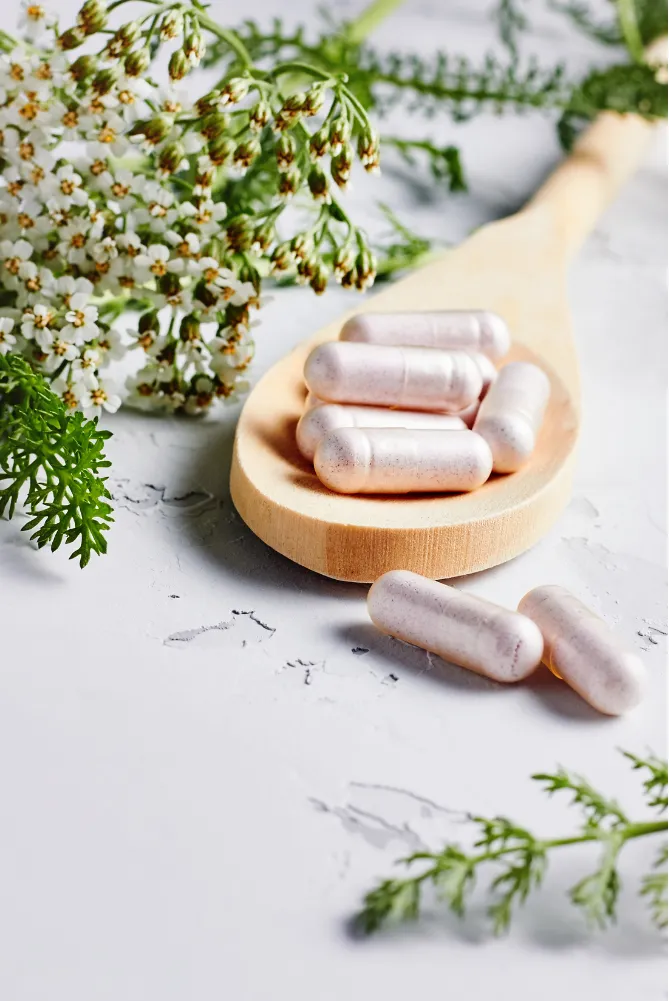 Herbal capsules on a wooden spoon with white flowers and green leaves on a textured surface.