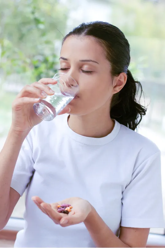 Person in a white shirt drinking water, holding colorful pills in hand, with a blurred green background.