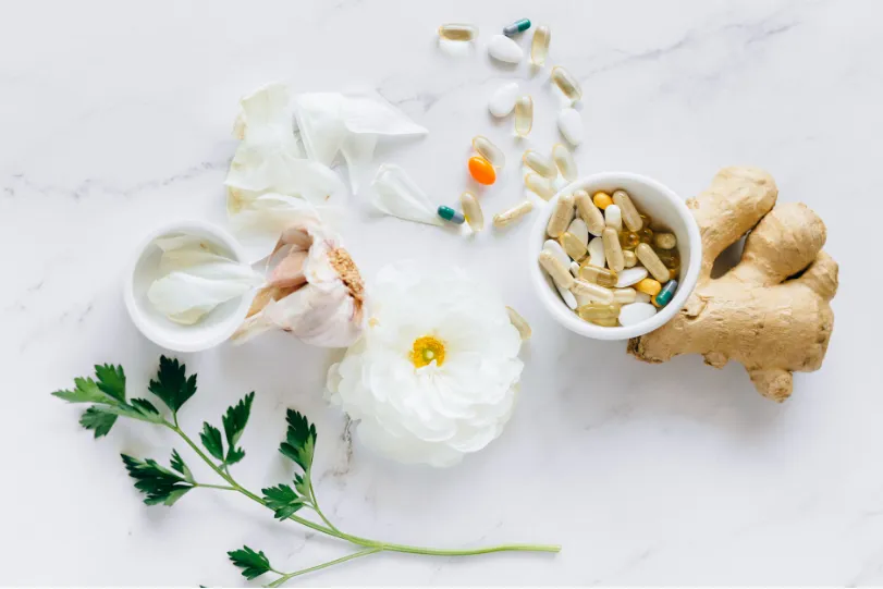 Herbs, garlic, ginger, pills, and a white flower on a white marble surface.