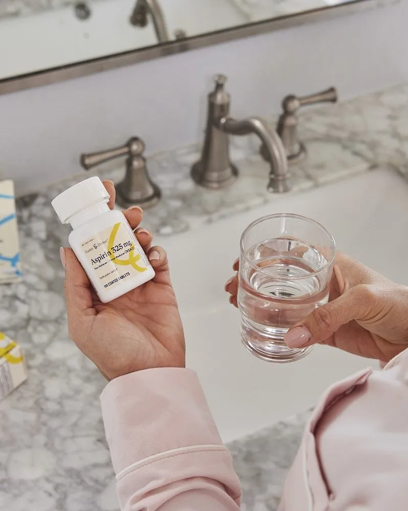 ft_shot11_med-product_006-5mb_1440x1000 1 Person holding a pill bottle in one hand and a glass of water in the other by a bathroom sink.